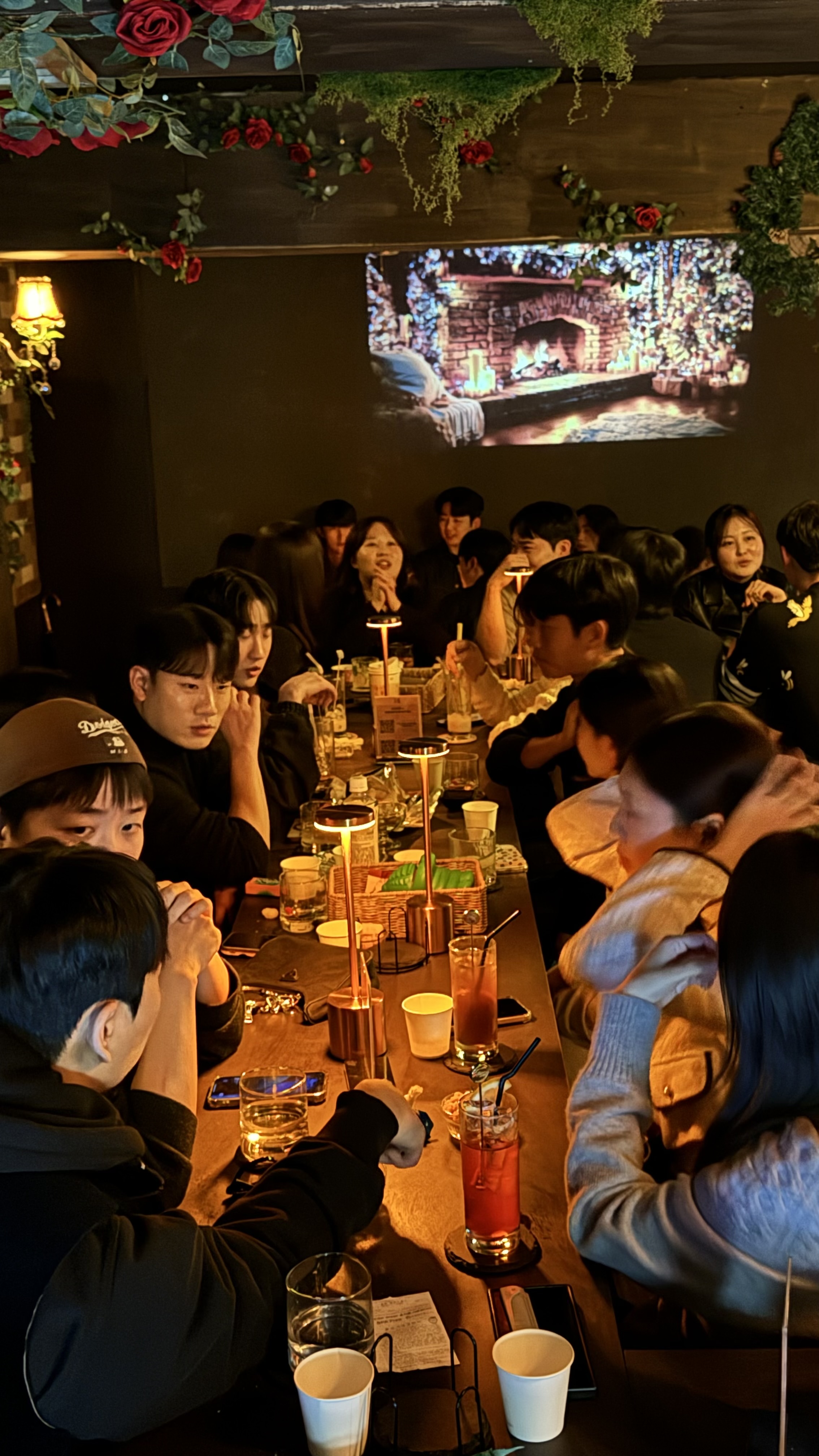장미와 그린 식물로 장식된 아늑한 분위기의 실내 매장 전경으로, 벽난로 영상이 재생되는 스크린 아래 긴 테이블에 손님들이 둘러앉아 칵테일과 음료를 즐기고 있다.