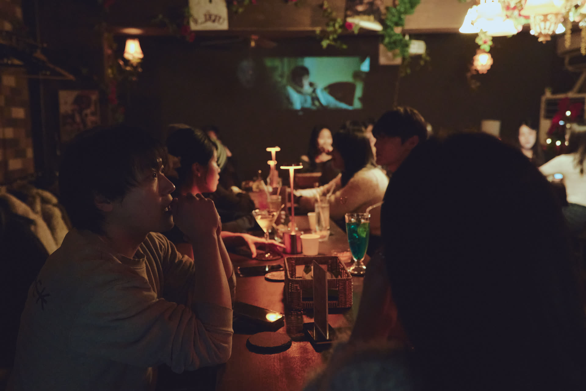 따뜻한 조명과 빈티지 소품으로 꾸며진 바(Bar) 인테리어 사진으로, 손님들이 긴 테이블에 앉아 칵테일을 즐기며 대화를 나누는 모습이 담겨 있다.