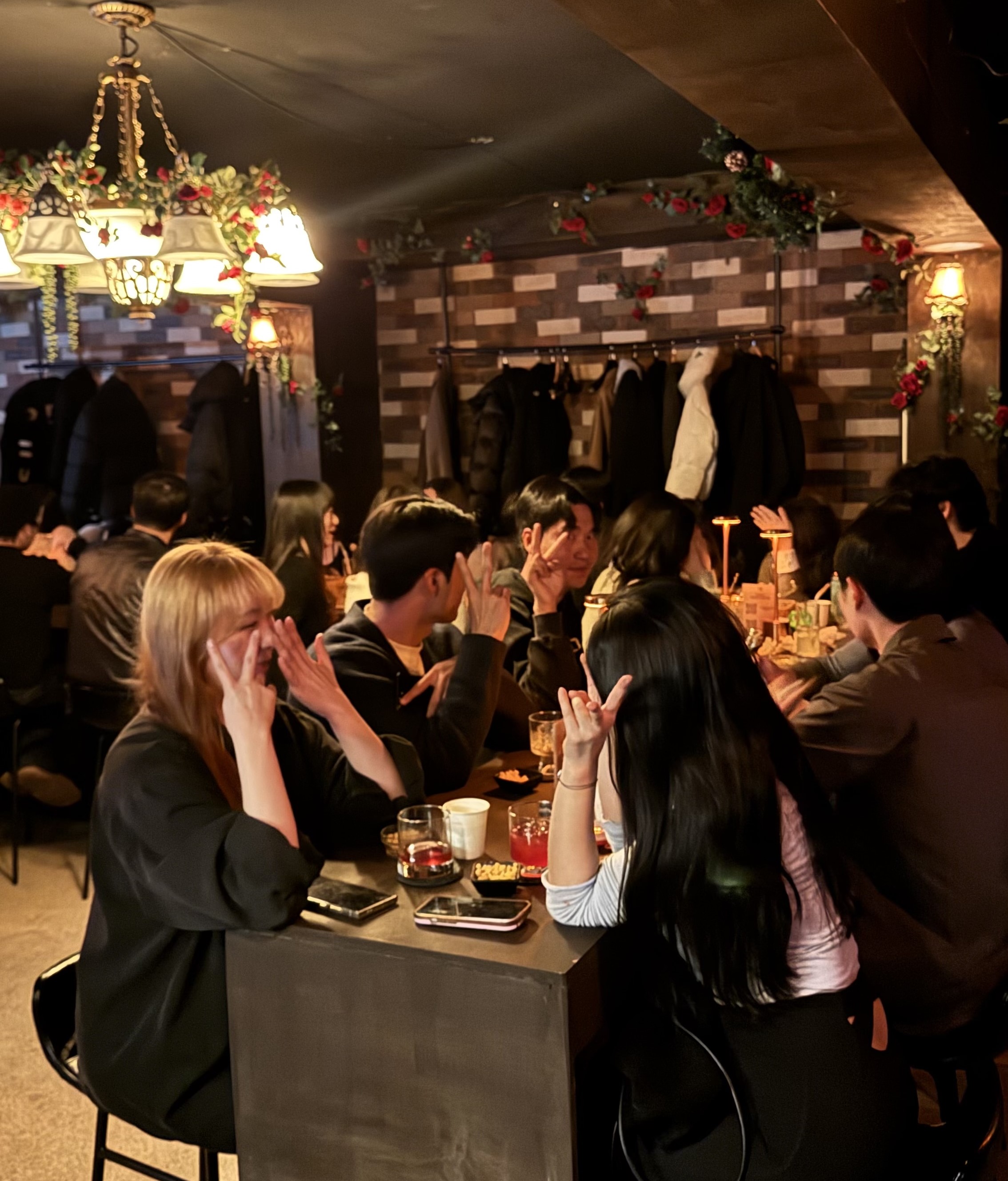 장미와 조명으로 꾸며진 아늑한 인테리어의 바에서 손님들이 테이블에 둘러앉아 음료를 즐기며 손 하트 포즈를 취하고 있는 모습으로, 매장 분위기와 손님 이용 장면을 보여주는 사진이다.
