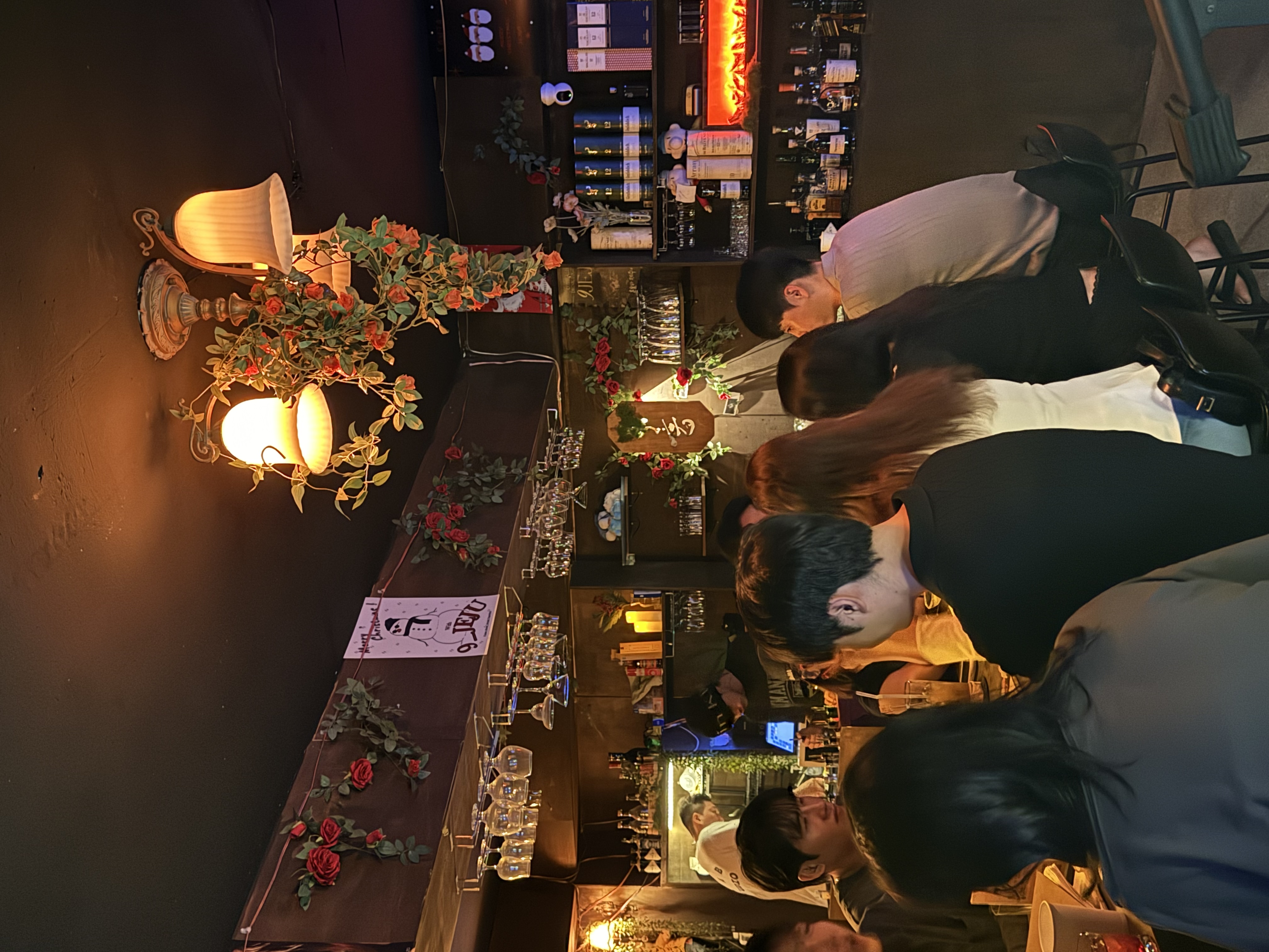 위스키·양주병이 진열된 바(bar) 인테리어 사진으로, 장미 장식과 조명 아래 다양한 술병 진열대가 보이고 손님들이 바 테이블에 모여 있는 모습이 담겨 있다.