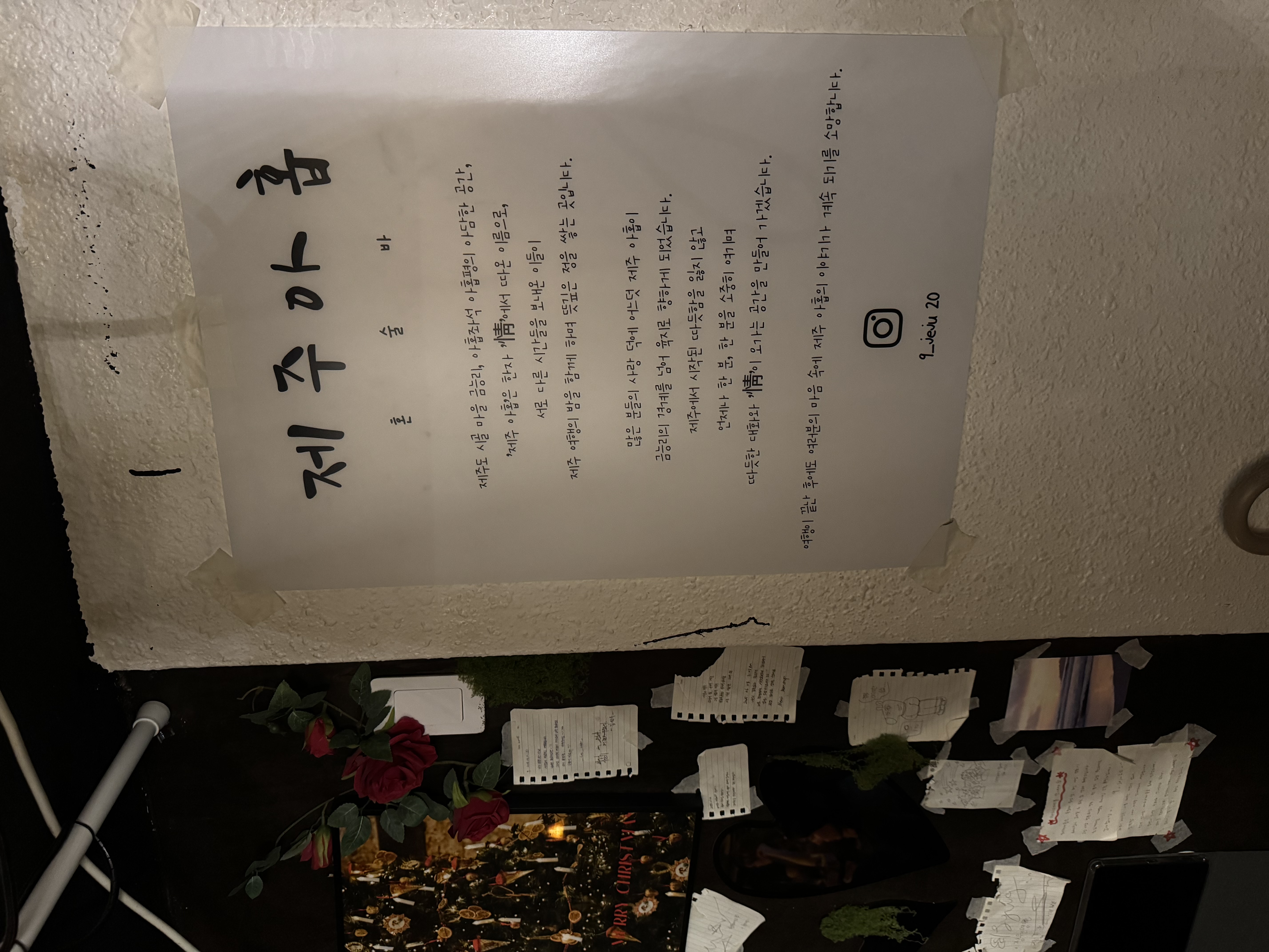 '제주마을'의 소개글이 적힌 벽면 포스터 사진으로, 아래쪽에는 손님들이 남긴 메모지와 낙서, 조화 장미, 크리스마스 카드 등으로 꾸며진 인테리어 소품들이 보인다.