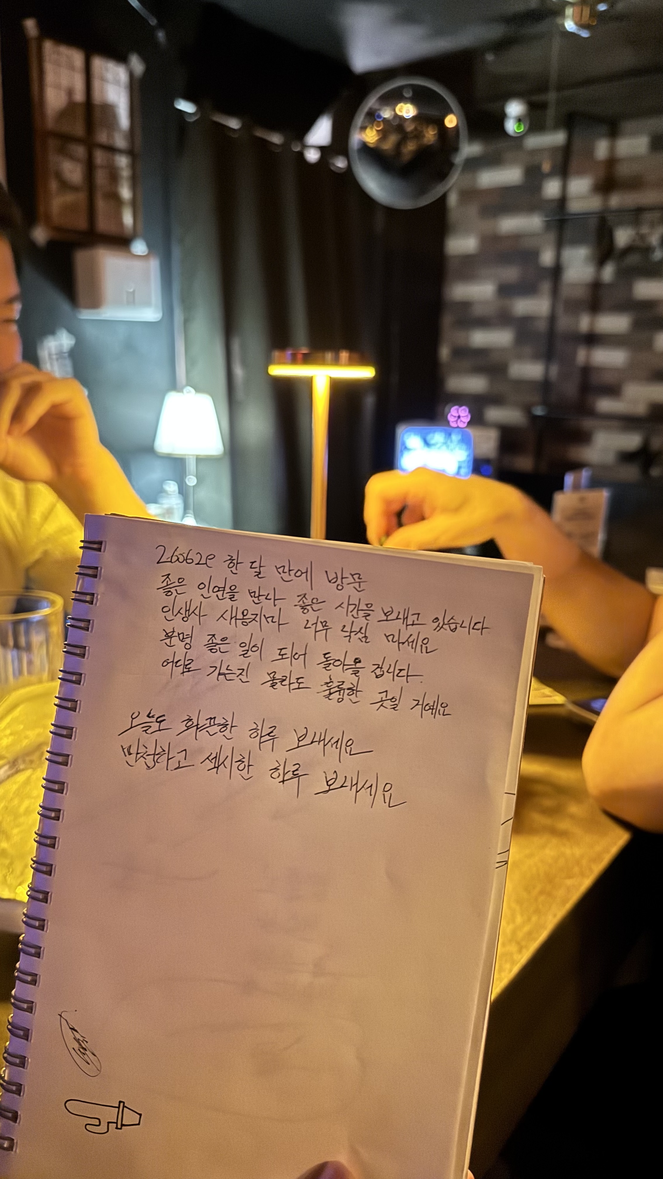 손님이 방명록 노트에 손글씨로 재방문 소감과 응원 메시지를 남긴 모습으로, 배경에는 조명이 은은한 매장 내부 인테리어와 테이블에 앉은 손님들이 보인다.