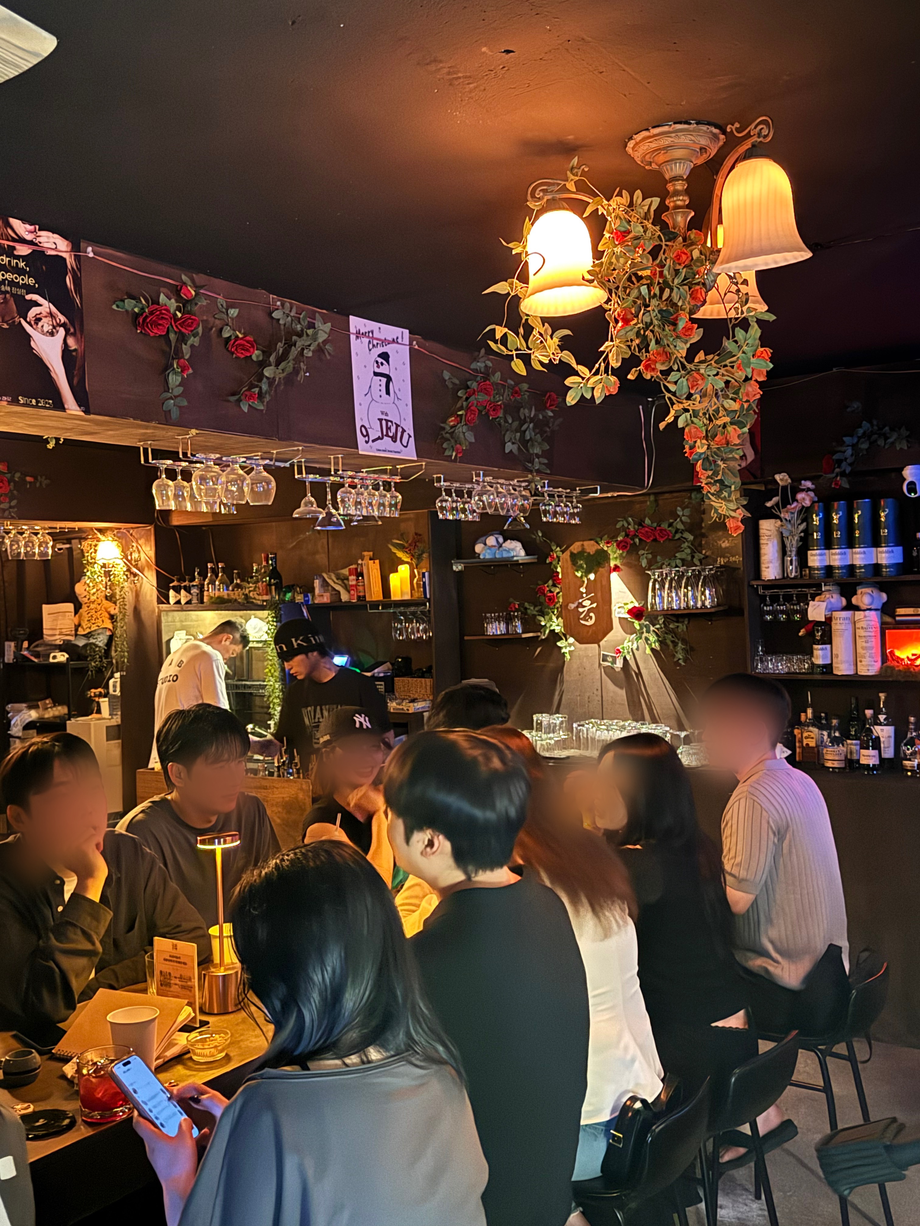 장미 넝쿨과 앤틱 조명으로 꾸며진 아늑한 분위기의 바 인테리어로, 바텐더가 음료를 준비하는 동안 많은 손님들이 테이블과 바 좌석을 가득 채우고 있는 활기찬 모습이 담긴 사진이다.