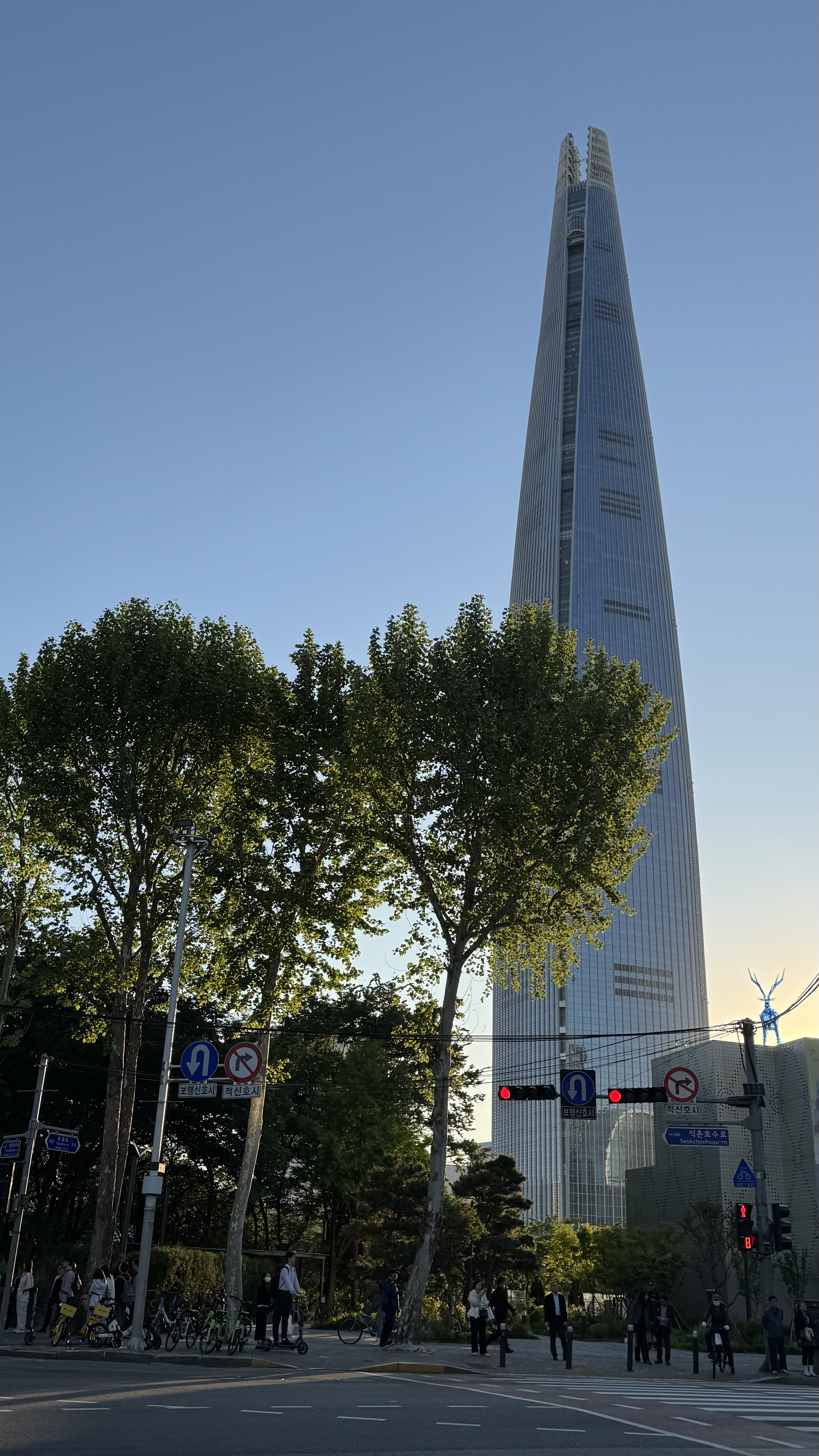 롯데월드타워가 우뚝 솟은 석촌호수 인근 거리 전경. 맑은 하늘과 가로수, 오가는 사람들이 어우러진 매장 위치를 짐작케 하는 랜드마크 사진.