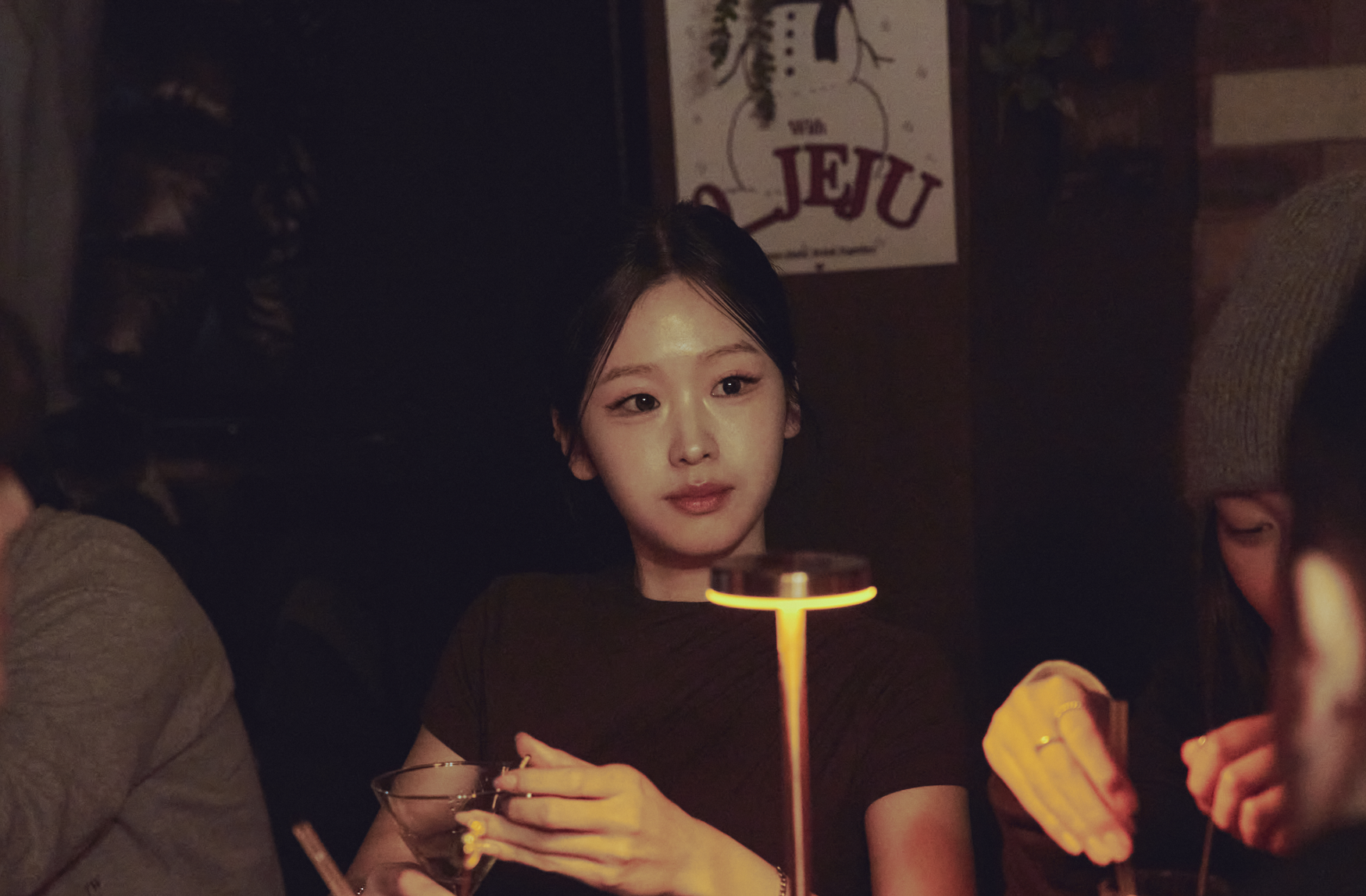 따뜻한 조명 아래 여성 손님이 칵테일 잔을 들고 앉아 있는 모습으로, 뒤편 벽에 'JEJU' 포스터가 붙어 있어 아늑한 바 분위기를 보여주는 인물 스냅컷이다.