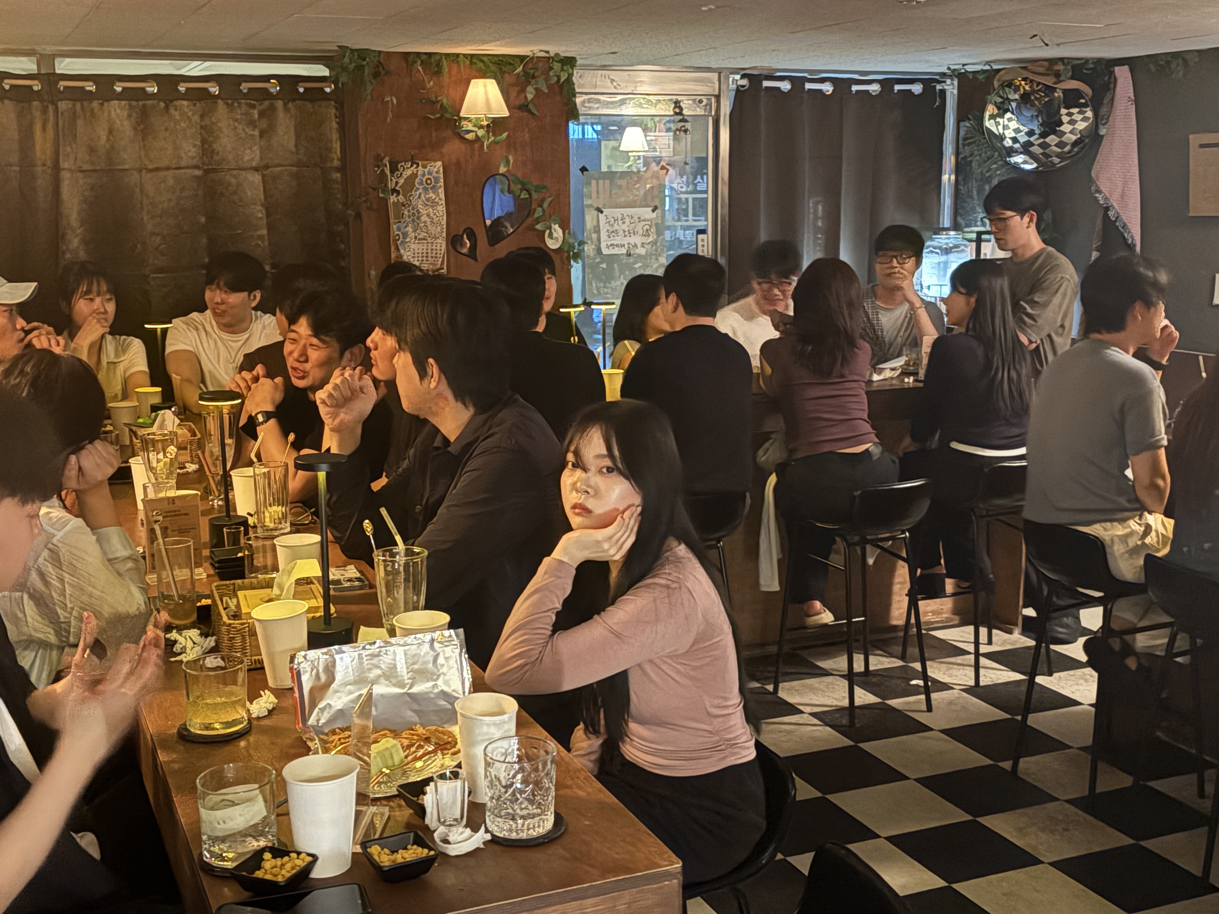 체크무늬 바닥과 아늑한 조명의 매장 내부 전경으로, 여러 테이블과 바 좌석에 손님들이 가득 앉아 있는 활기찬 분위기를 담았다.
