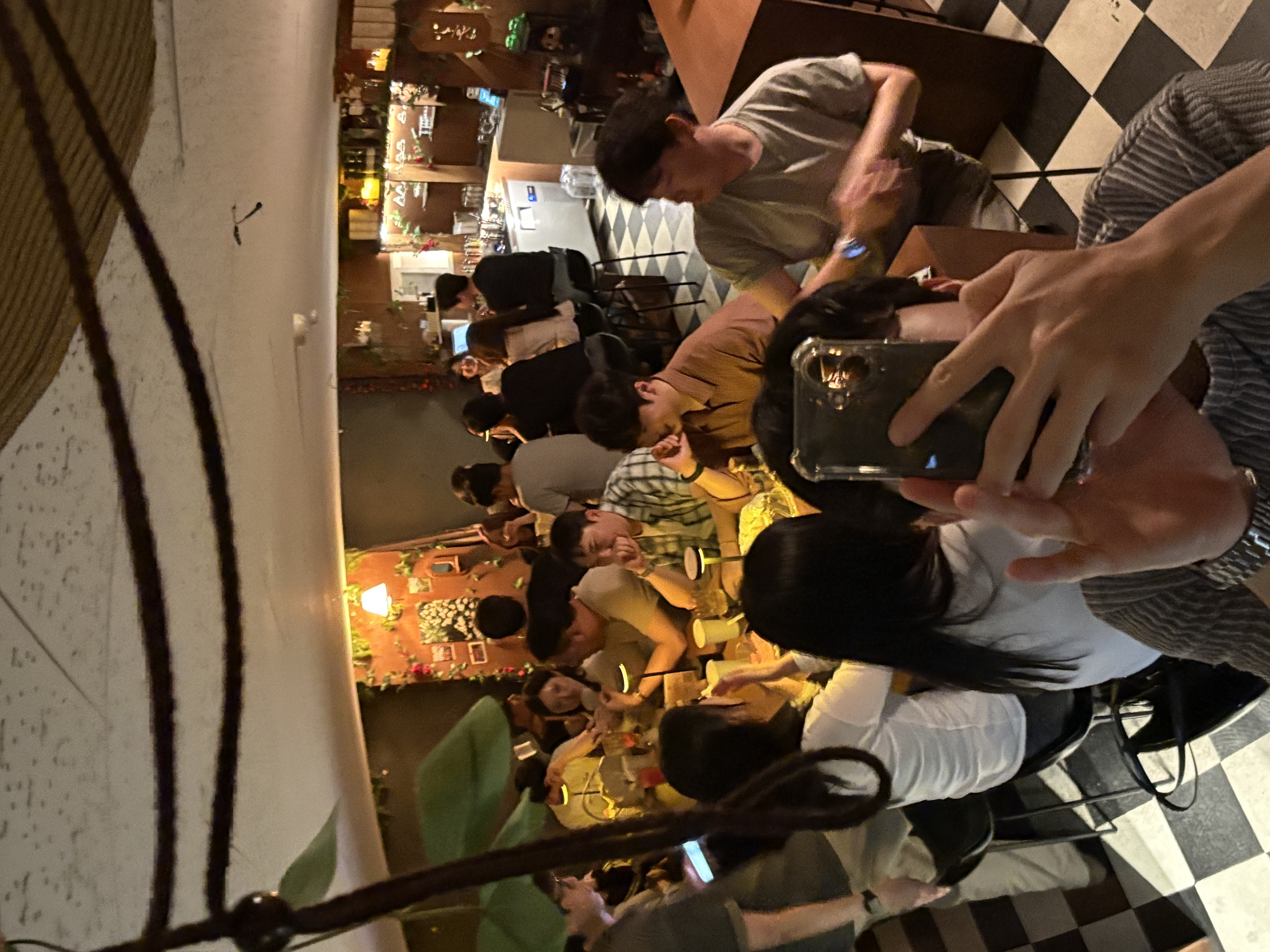 체크무늬 바닥의 홀에서 손님들이 테이블에 둘러앉아 대화하고 사진을 찍는 모습으로, 매장의 활기찬 분위기를 보여주는 단체 손님 사진.