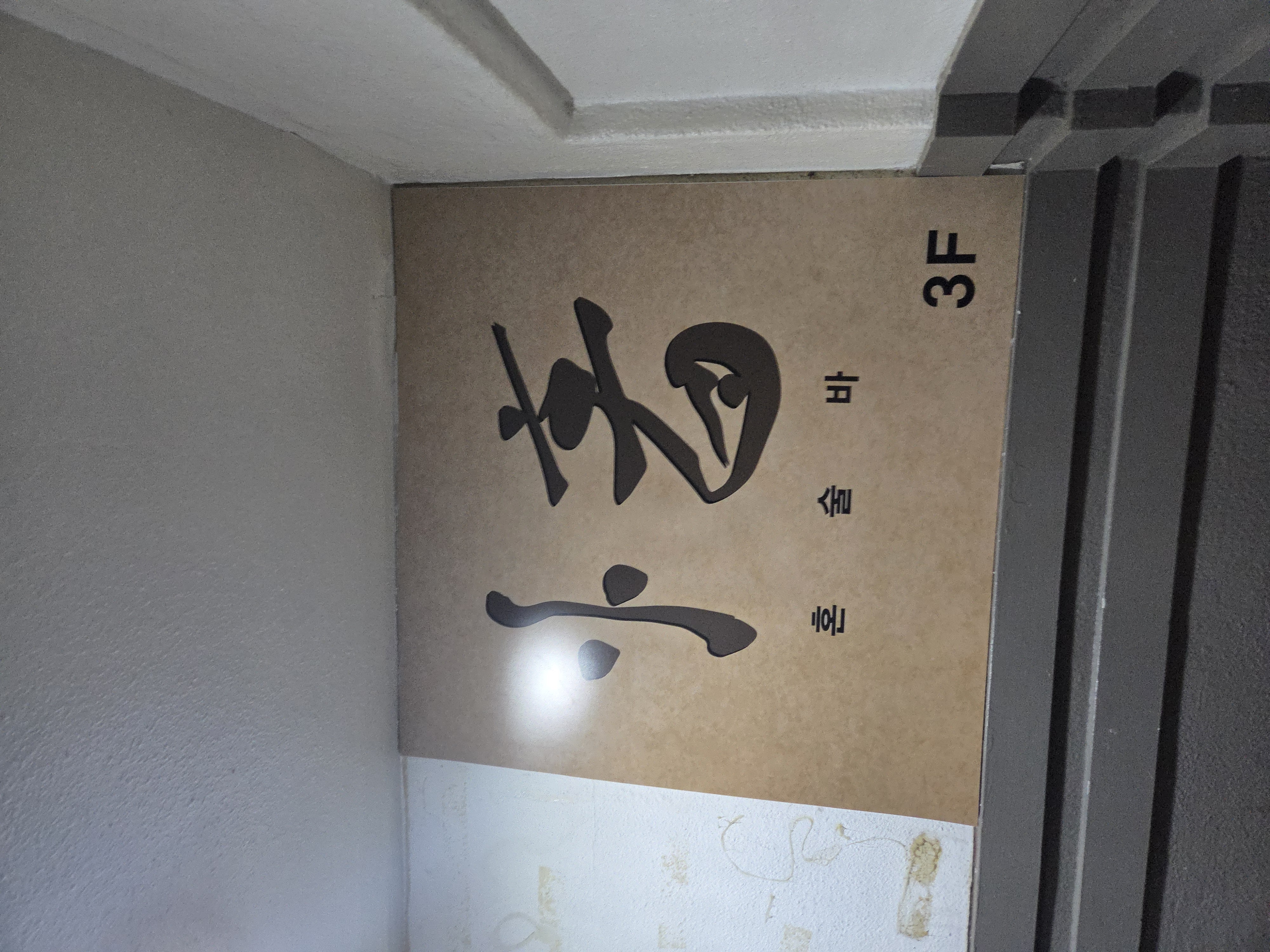 간판 클로즈업 사진 - 계단 입구 벽면에 우드톤 바탕의 캘리그래피 로고와 '3F' 층수 표시가 부착되어 있다.
