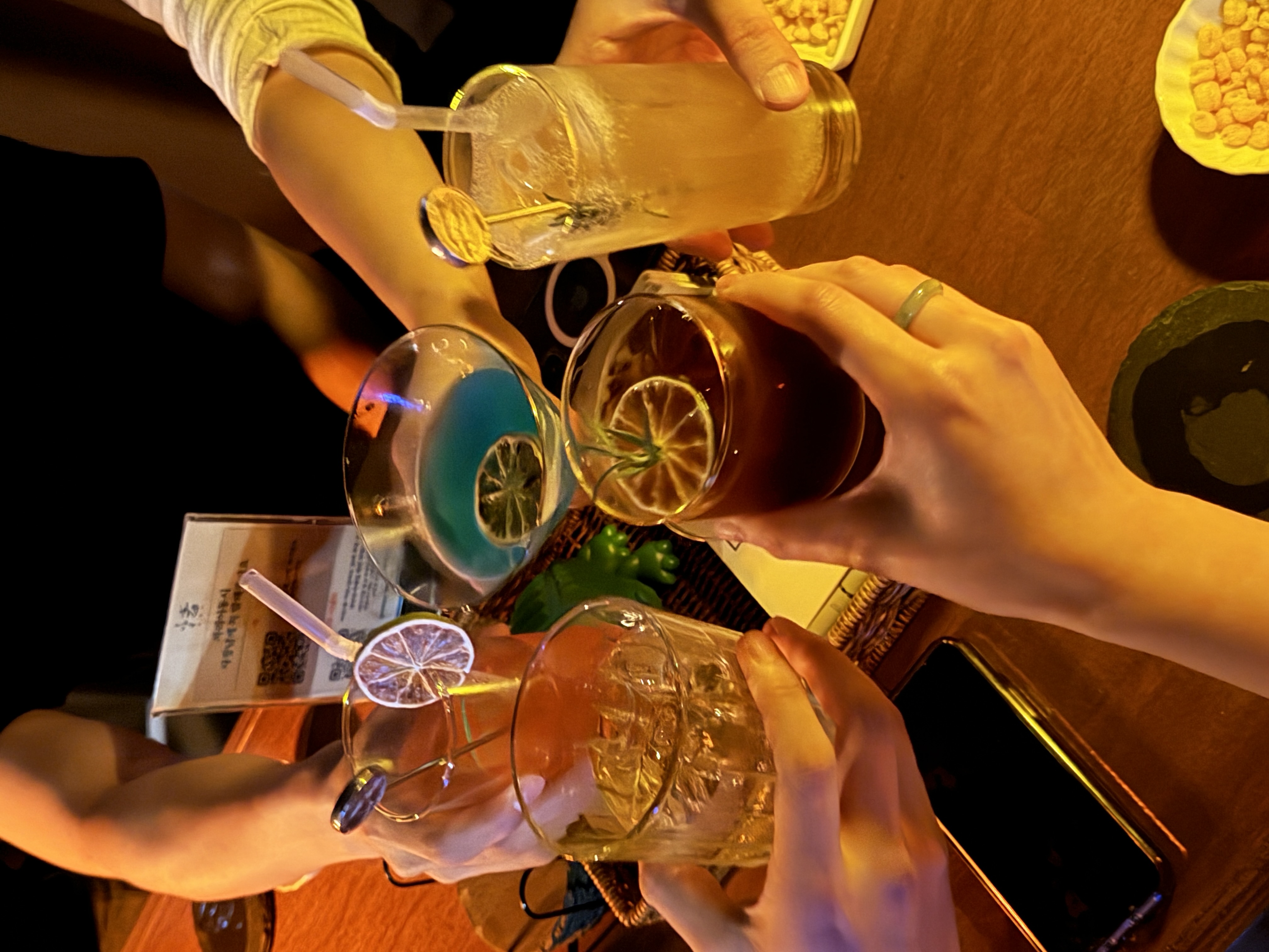 칵테일/음료 건배 사진. 라임 슬라이스가 올라간 다양한 색깔의 칵테일 잔(블루, 앰버, 클리어 톤)을 여러 사람이 함께 부딪히는 장면으로 메뉴와 분위기를 동시에 보여줌.