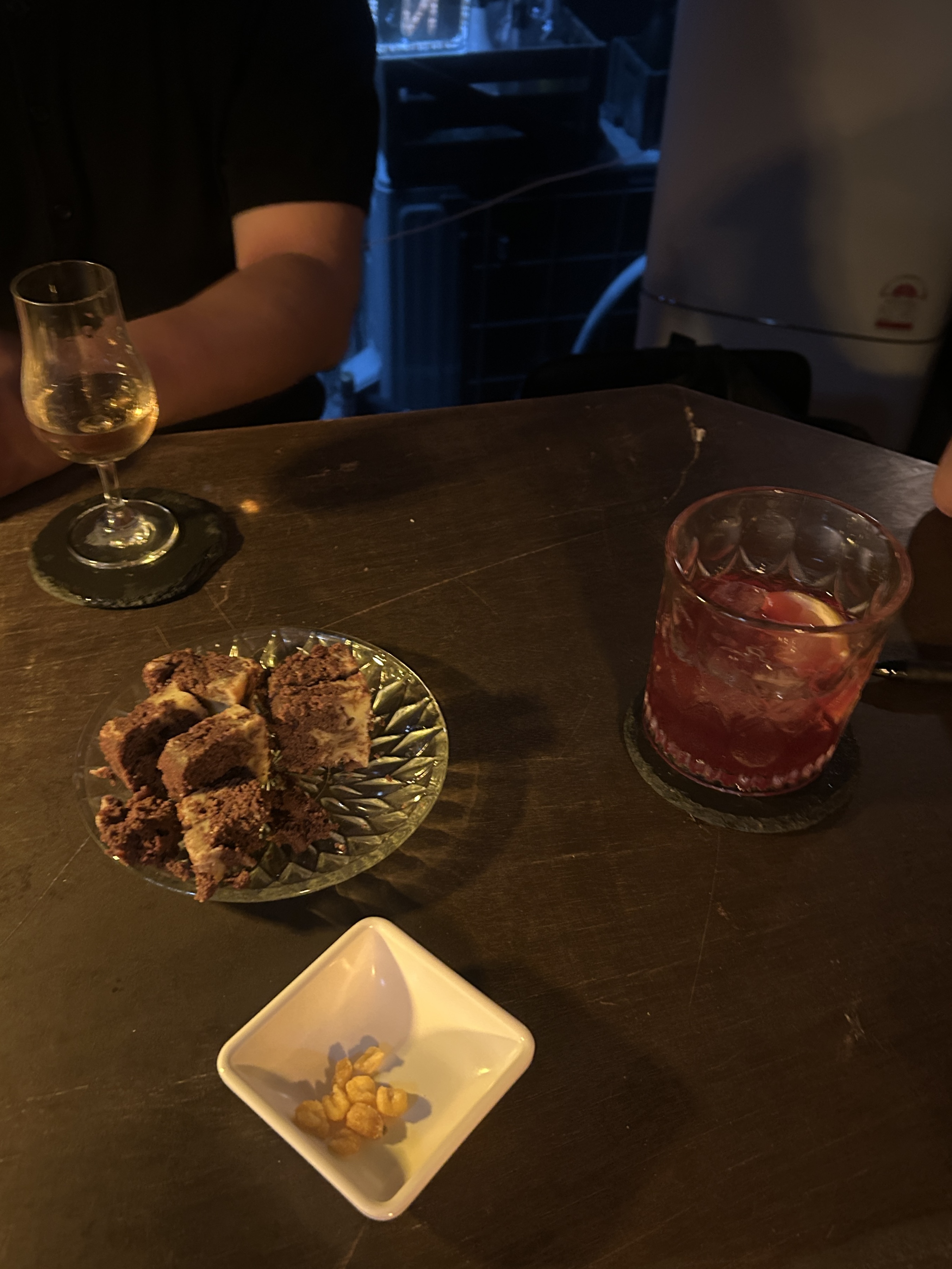 바 테이블 위 위스키 한 잔과 붉은빛 칵테일, 초코 크럼블 치즈케이크 조각, 견과류 안주가 함께 놓인 술상 사진.